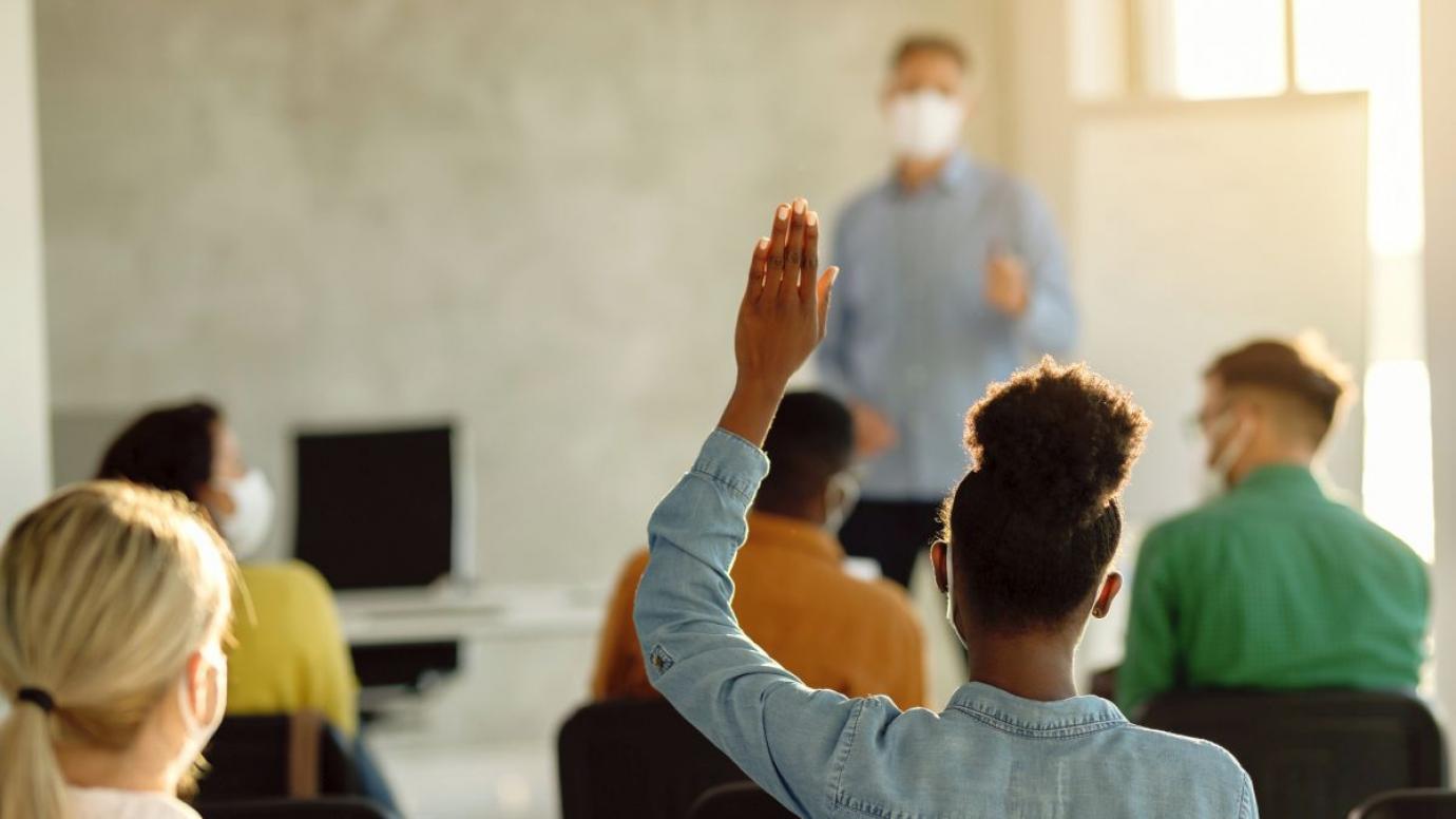 Back view of black female student raising arm to ask a question during a lecture in the classroom.